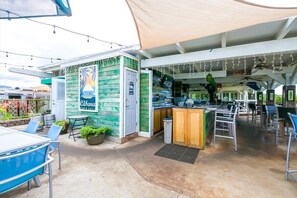 An outdoor seating area with a bar and grill, tables, chairs, and a canopy providing shade. - Stunning Ocean View Condo on the beach w AC, Kiahuna Plantation, Poipu Beach (Koloa)