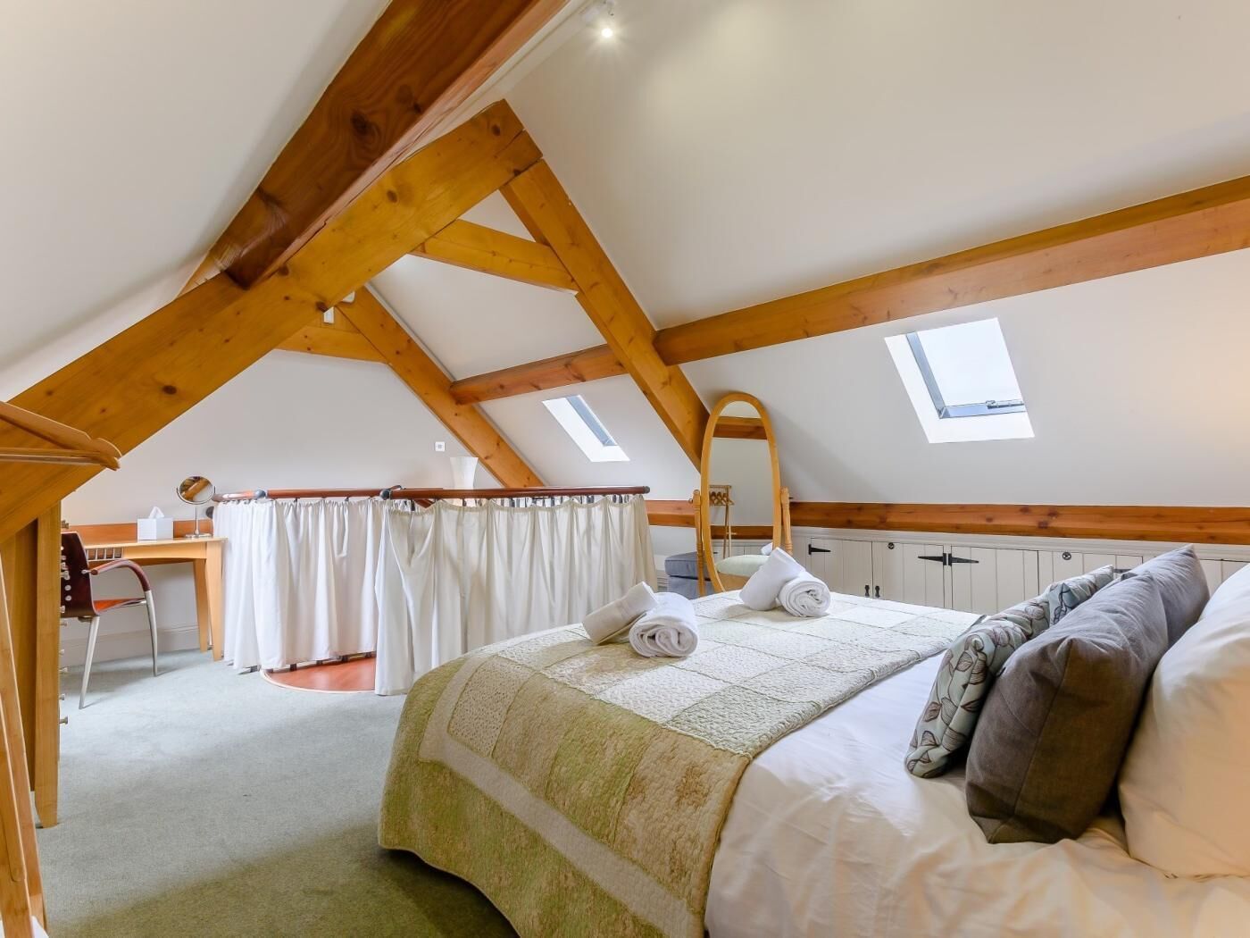 A bedroom with a sloped ceiling, wooden beams, a bed with pillows, a desk, and a chair.