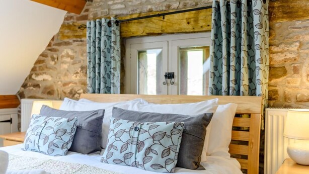 A bedroom with a stone wall, a wooden bed frame, patterned pillows, and a window with curtains. - 2 Bed in Crickhowell (oc-42920) (Llangattock)