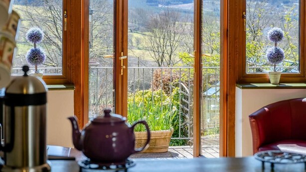 Um quarto com vista para a paisagem campestre através de grandes janelas, uma cadeira vermelha, uma mesa com uma chaleira e uma planta em vaso no parapeito da janela.