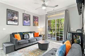 A living room with a grey sofa, a coffee table, and a ceiling fan. There are framed posters on the wall and a view of greenery outside. - Galactic Oasis in Kissimmee (Davenport)