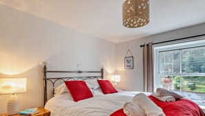A bedroom with a metal bed frame, white bedding, red pillows, a bedside lamp, a window with a view, and a hanging light fixture. - 3 Bed in Minehead (oc-44078) (Porlock)
