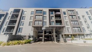 A modern multi-story building with a central entrance, balconies, and a clear sky. - Cozy 1-bedroom apartment in superb Pretoria with gym, pool (Pretoria)
