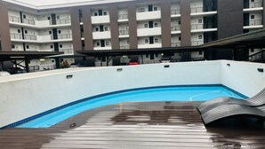 A swimming pool with a wooden deck, a covered area, and a multi-story building in the background. - Cozy 1-bedroom apartment in superb Pretoria with gym, pool (Pretoria)