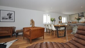 Una sala moderna con un sofá de cuero marrón, un comedor con mesa y sillas de madera, y una cocina al fondo.