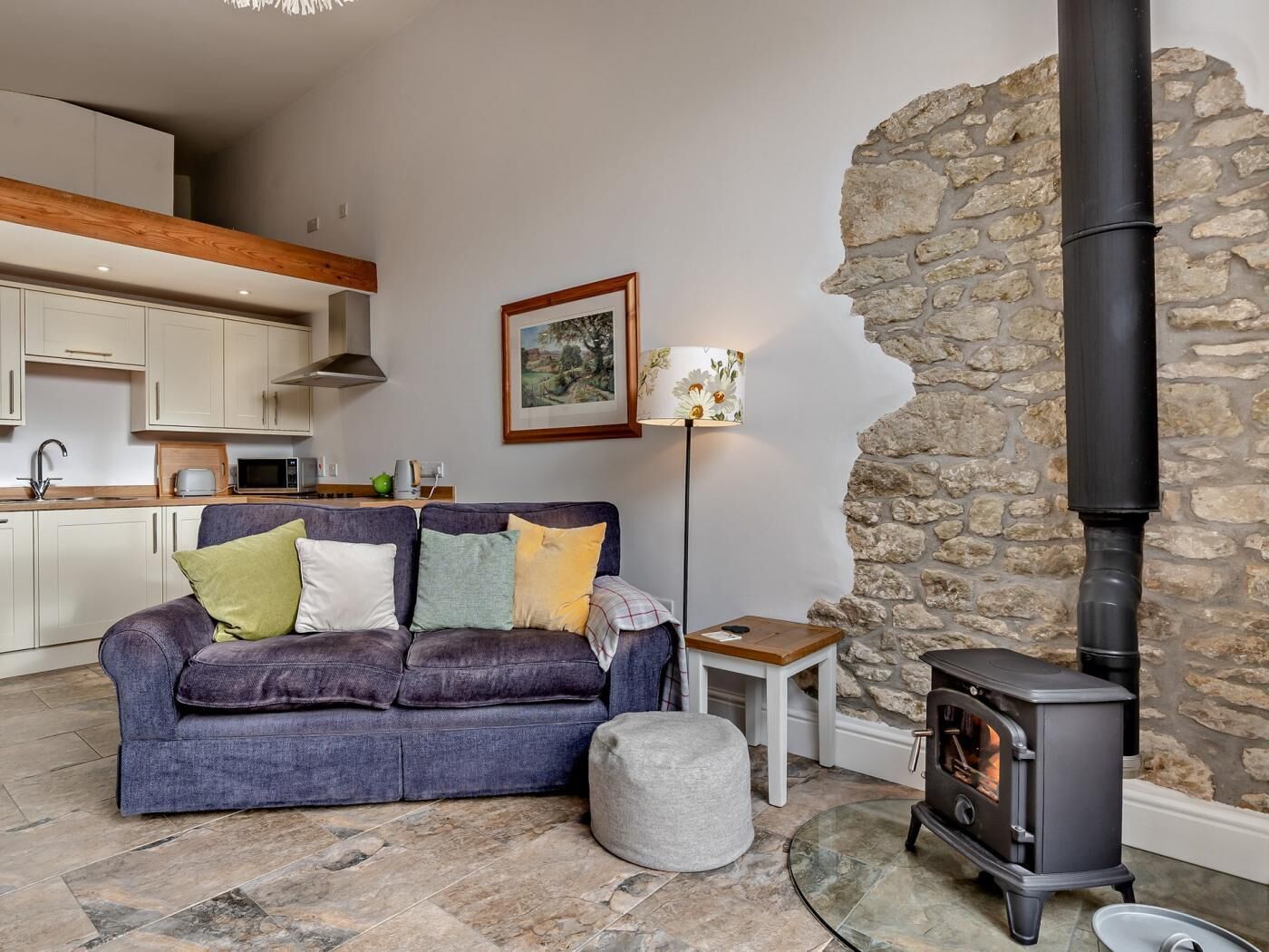 A cozy living room with a sofa, a small table, a lamp, a stone wall, and a wood-burning stove.