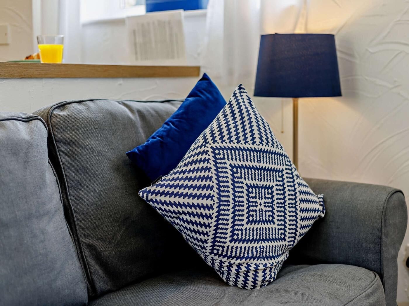A grey sofa with a blue and white patterned pillow and a dark blue lamp.