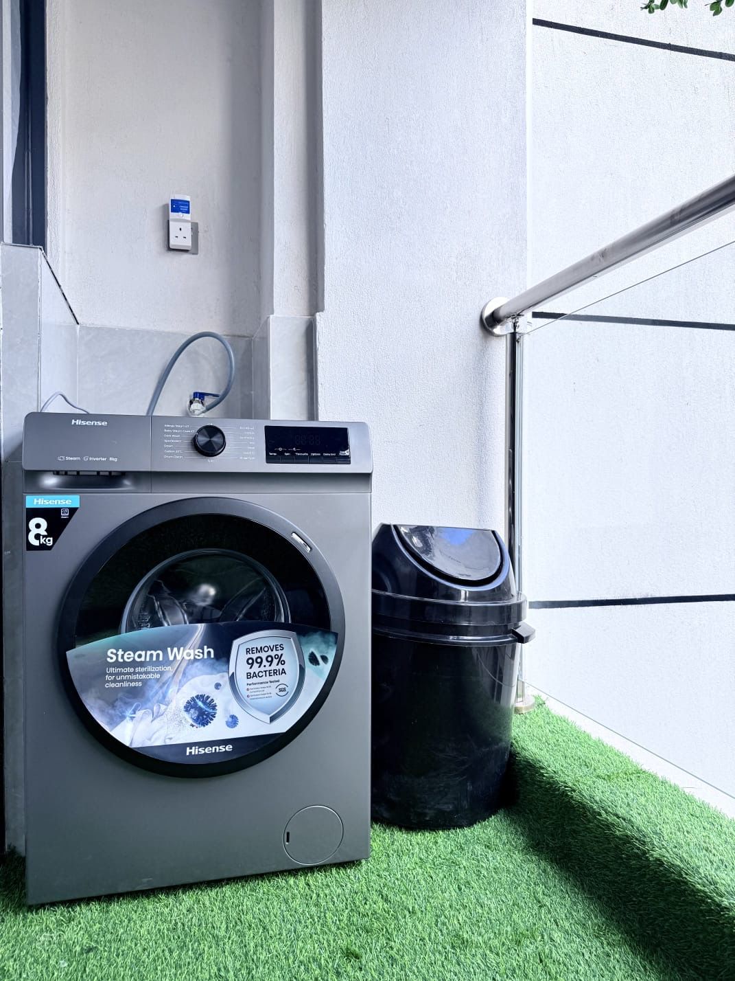 Eine Hisense-Dampfwaschmaschine mit digitaler Anzeige, auf künstlichem Rasen neben einem schwarzen Mülleimer und einer weißen Wand platziert.
