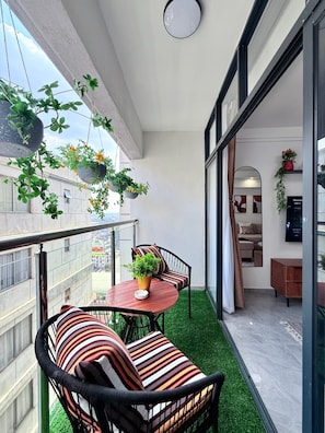 Un balcón con césped artificial, una mesa redonda, dos sillas acolchadas, plantas colgantes y vistas de la ciudad.