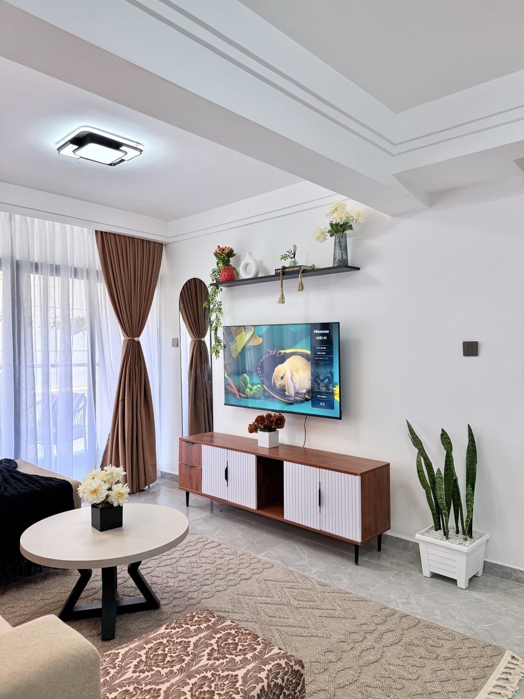 Een moderne woonkamer met een flatscreen televisie, een ronde salontafel, een bank, een gestreept voetenbankje, een potplant en een decoratieve plank met bloemen.