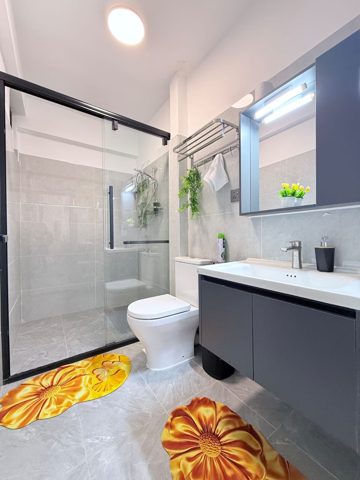 Un baño moderno con cabina de ducha de vidrio, un inodoro, un lavabo con espejo y felpudos decorativos con motivos florales en el suelo.