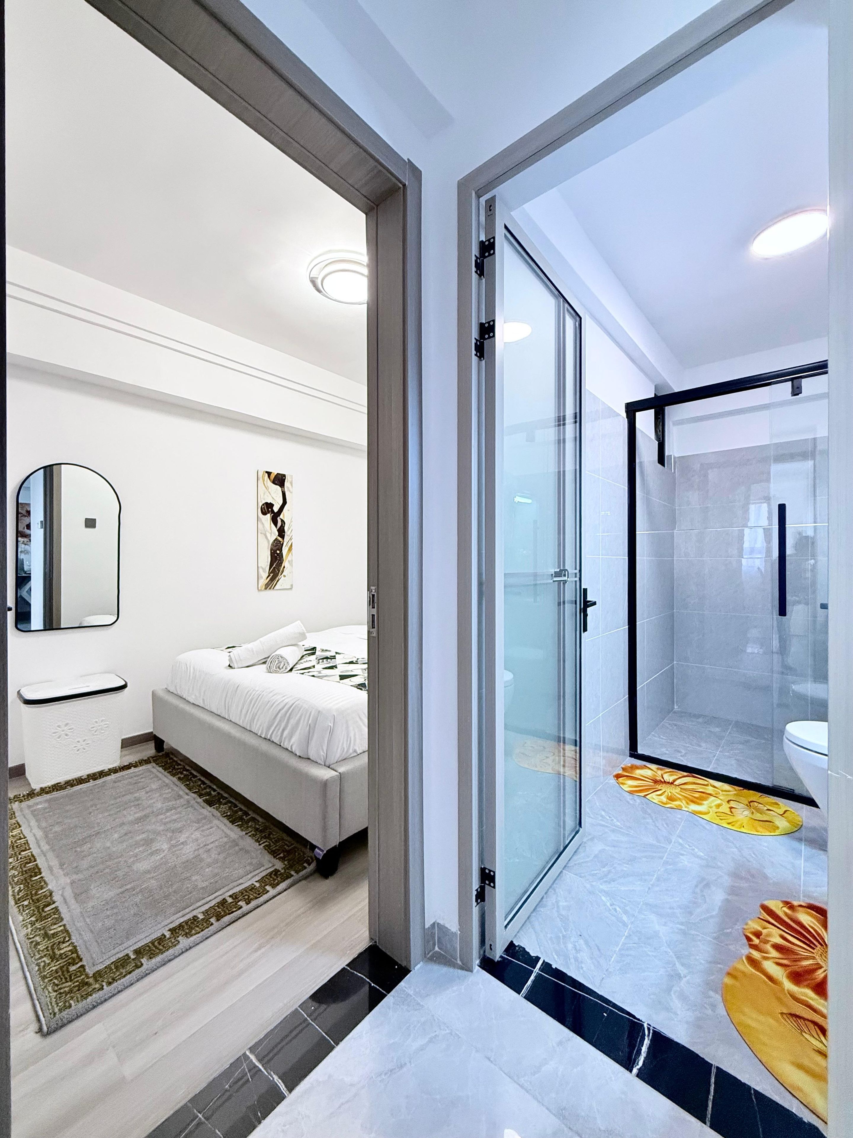 Ein modernes Hotelzimmer mit Bett, einem Badezimmer mit Glasdusche und einem Gang zu einem weiteren Zimmer.