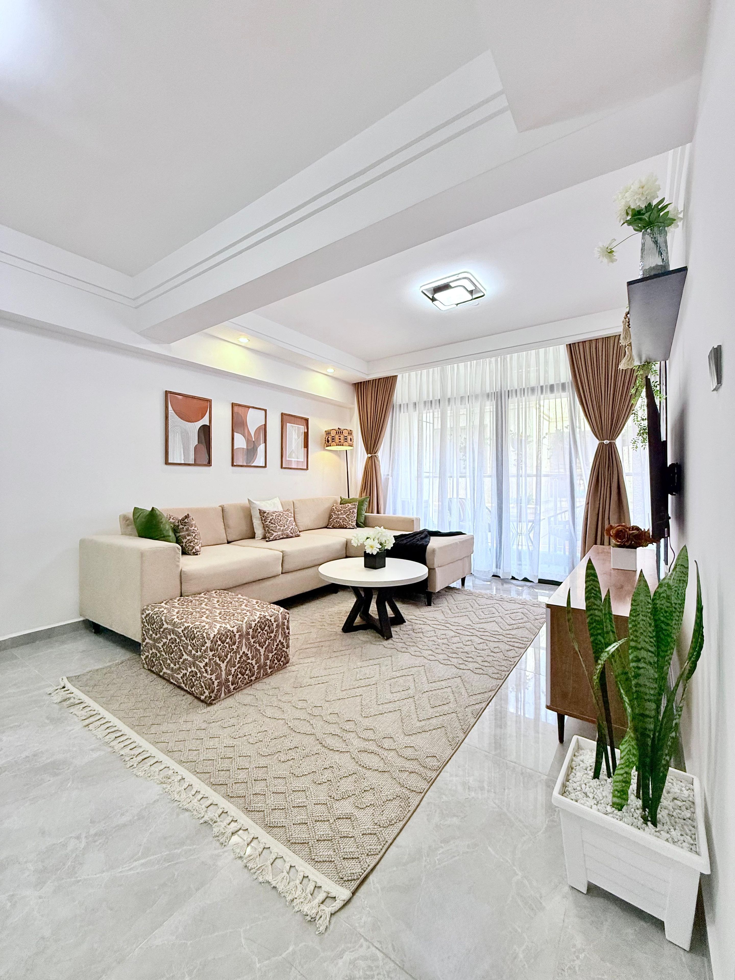 Una sala de estar moderna con un sofá color beige, un otomán estampado, una mesa de centro y un ventanal con cortinas.