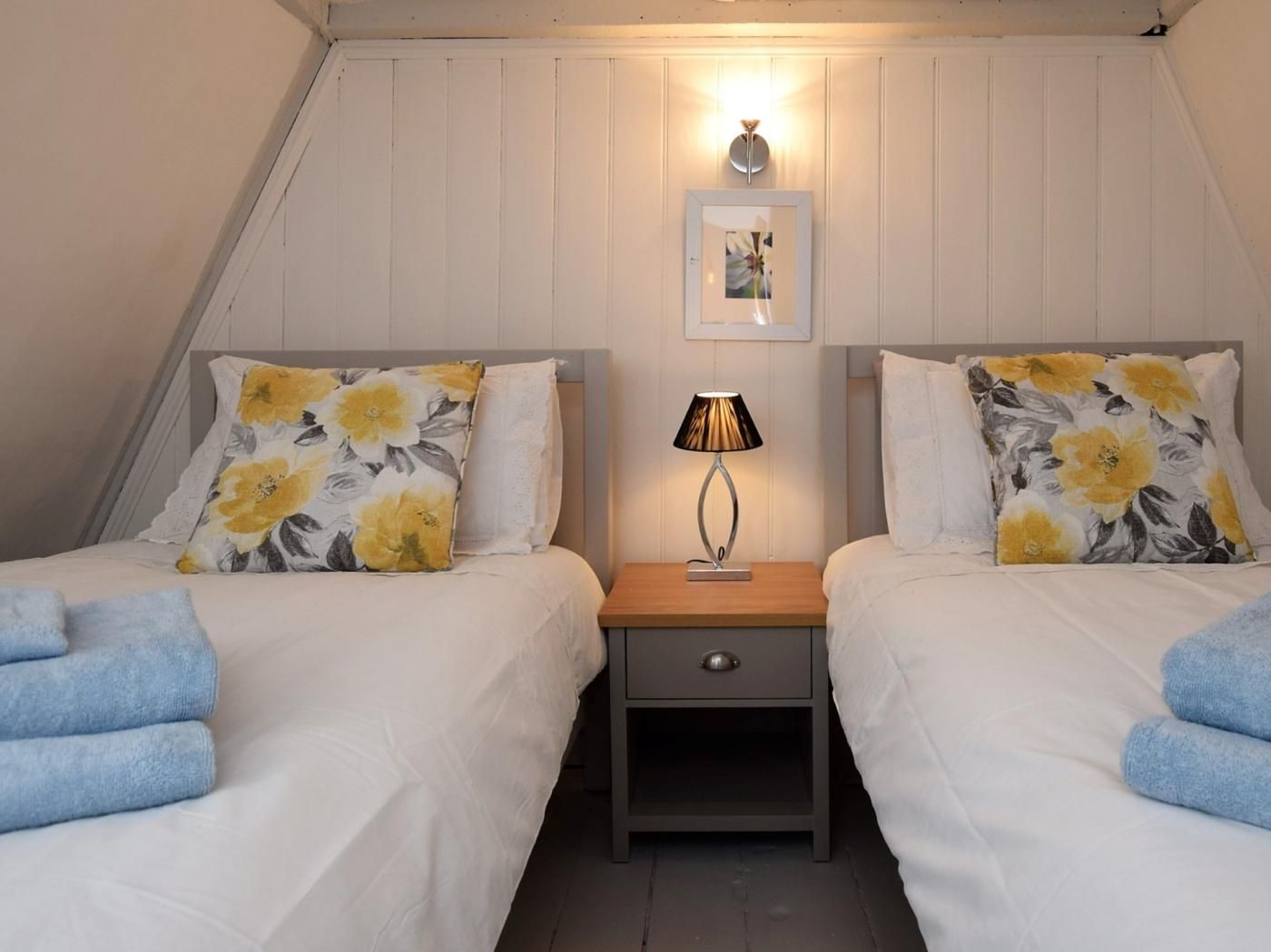 A bedroom with two beds, each with floral pillows, a bedside table with a lamp, and a framed picture on the wall.