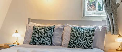 Una cama bien hecha con una toalla blanca enrollada, dos almohadas estampadas, una lámpara de pared, un cuadro enmarcado y una ventana con cortinas.