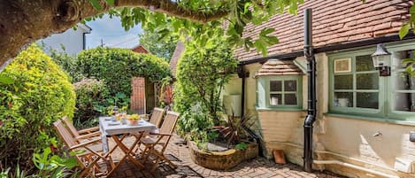 Eine Ziegelterrasse mit Tisch und Stühlen, umgeben von Grünpflanzen und einem Haus mit grünem Fenster.