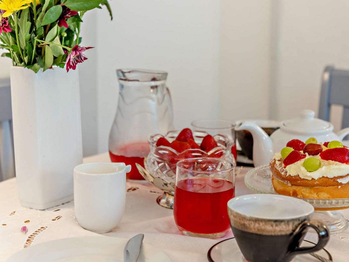 A table set with a pitcher, teapot, cups, a glass pitcher, and a dessert with fruit on a white tablecloth.