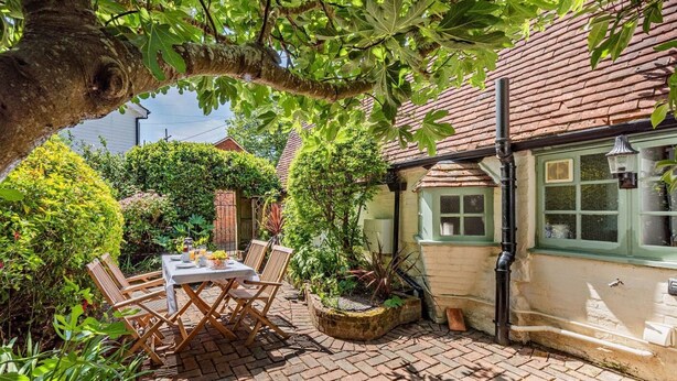 Eine Ziegelterrasse mit Tisch und Stühlen, umgeben von Grünpflanzen und einem Haus mit grünem Fenster.