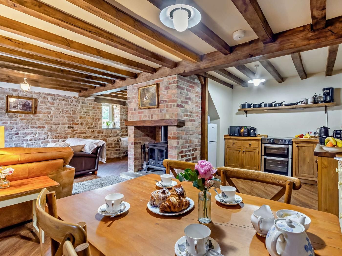 Un interior rústico con vigas de madera a la vista, una chimenea de ladrillo, una mesa de comedor dispuesta con tazas y pasteles, y una cocina con electrodomésticos modernos.