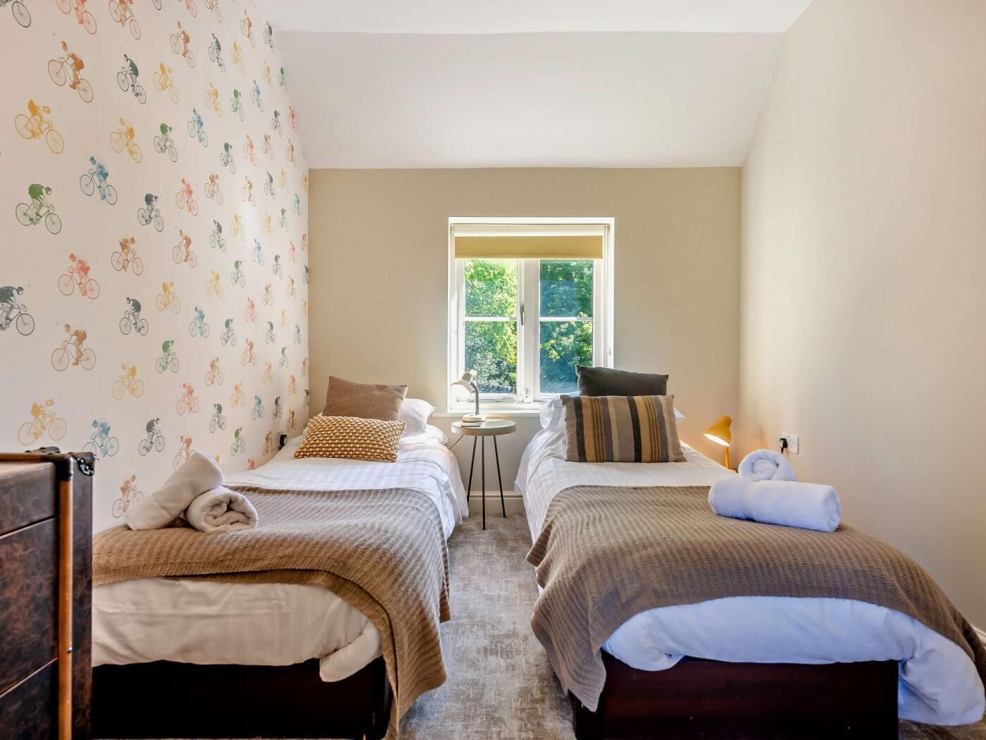 A room with two beds, a small table, a window with a view, and a patterned wall.