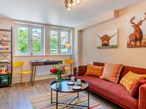 Ein gemütliches Wohnzimmer mit einem roten Sofa, einem kleinen runden Couchtisch, einem Bücherregal und einem Schreibtisch mit einer Lampe.