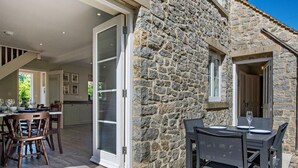 A stone building with a dining area, a kitchen visible through a glass door, and a patio with outdoor seating. - 4 Bed in Matlock (oc-47450) (Brassington)