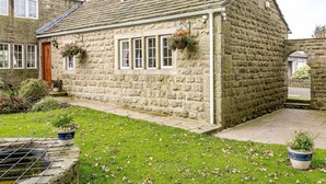 A stone cottage with a sloped roof, a wooden door, and a small garden with potted plants. - 1 Bed in Sheffield (oc-48259) (Thurlstone)