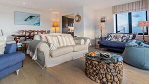 Ein modernes Wohnzimmer mit einem beigefarbenen Sofa, einem blauen Sessel, einem runden Couchtisch mit Holzscheiten und einem Essbereich mit einem Holztisch und Stühlen.