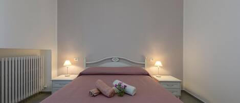 A neatly made bed with a pink bedspread, two bedside lamps, a white radiator, and a small table with a lamp.