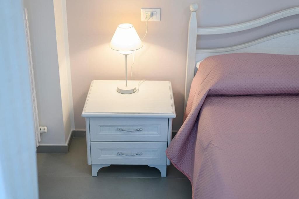 A white bedside table with a lamp, a pink bedspread, and a white headboard.