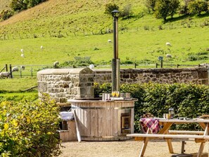 Un espace extérieur rustique aménagé avec un four en pierre, une table recouverte d’une couverture et une vue sur un champ verdoyant où paissent des moutons.