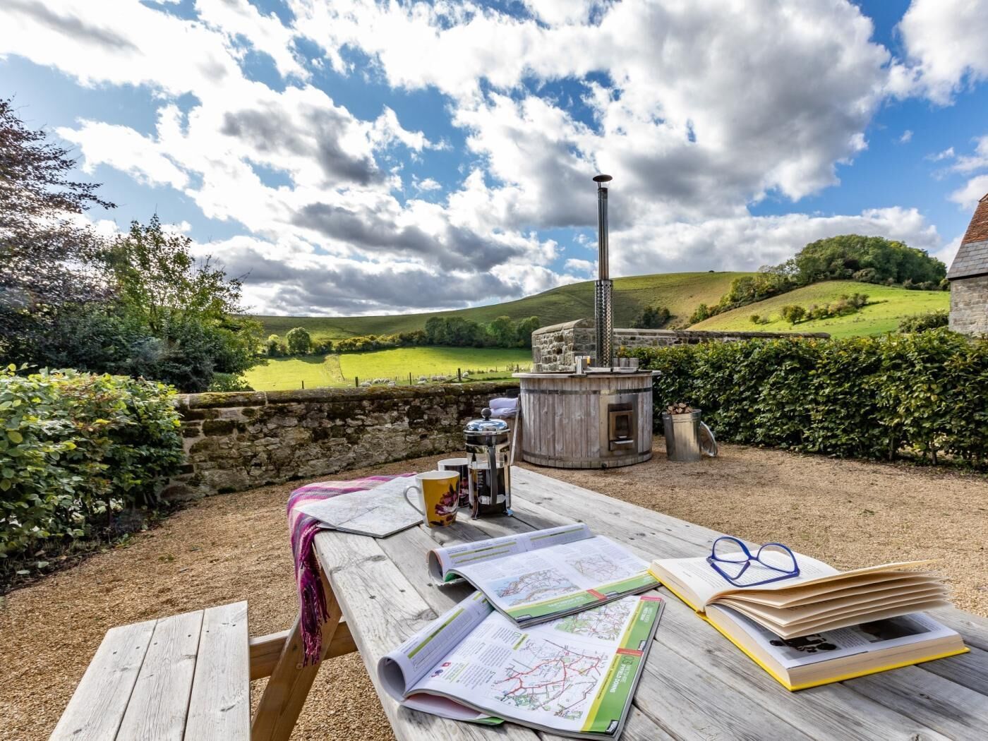 Una mesa de madera con mapas, un libro y gafas, al aire libre con un muro de piedra y vistas a colinas onduladas.