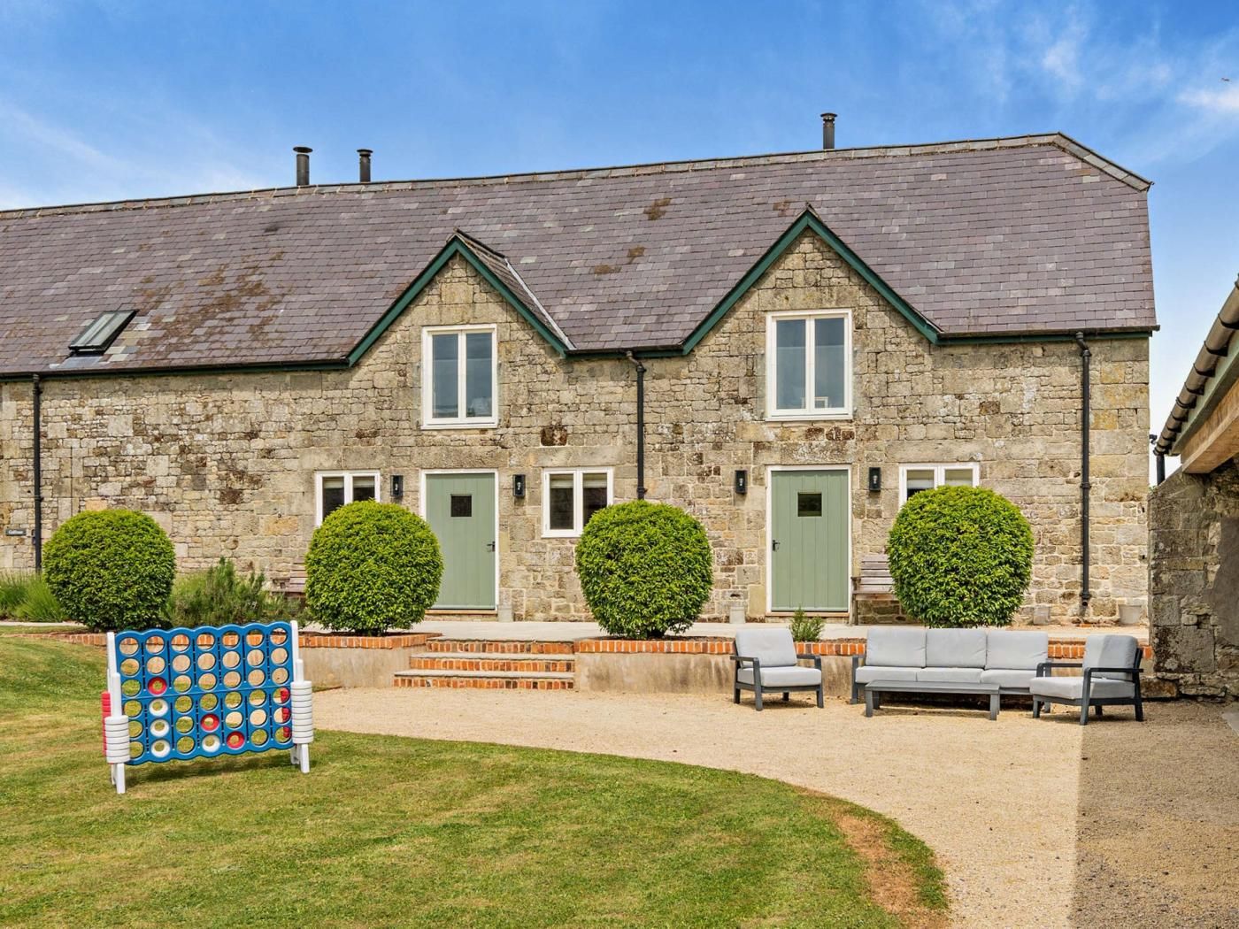 A stone building with a gabled roof, two windows on the ground floor, and a green door. There are three seating areas with chairs and a small table.