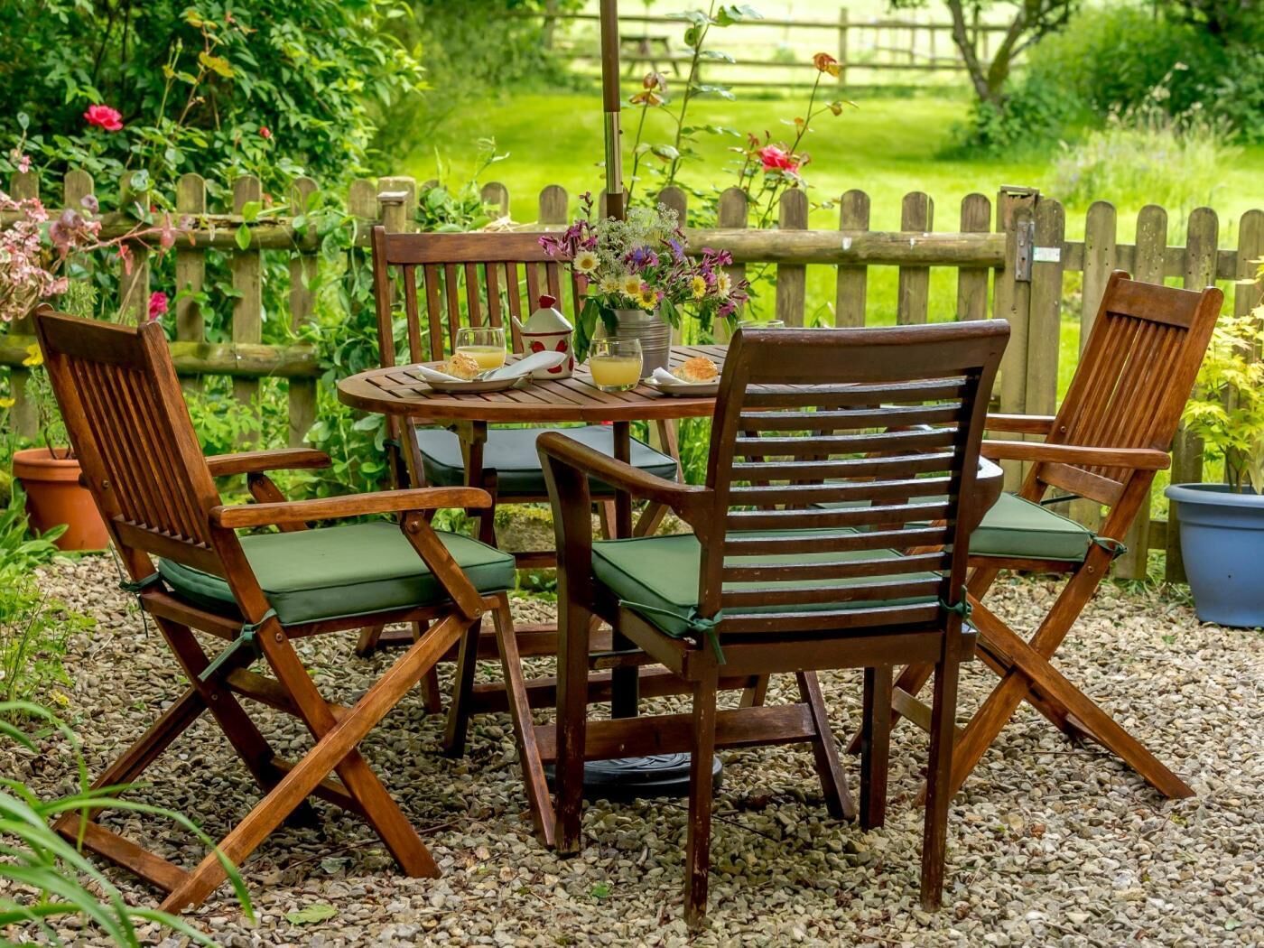 A wooden outdoor table with chairs, surrounded by gravel, with a wooden fence and greenery in the background.