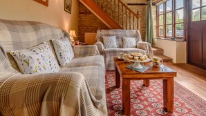 A cozy living room with a sofa, armchair, and a coffee table with pastries. A staircase with wooden railings is visible in the background. - 2 Bed in Cirencester (oc-48925) (Daglingworth)