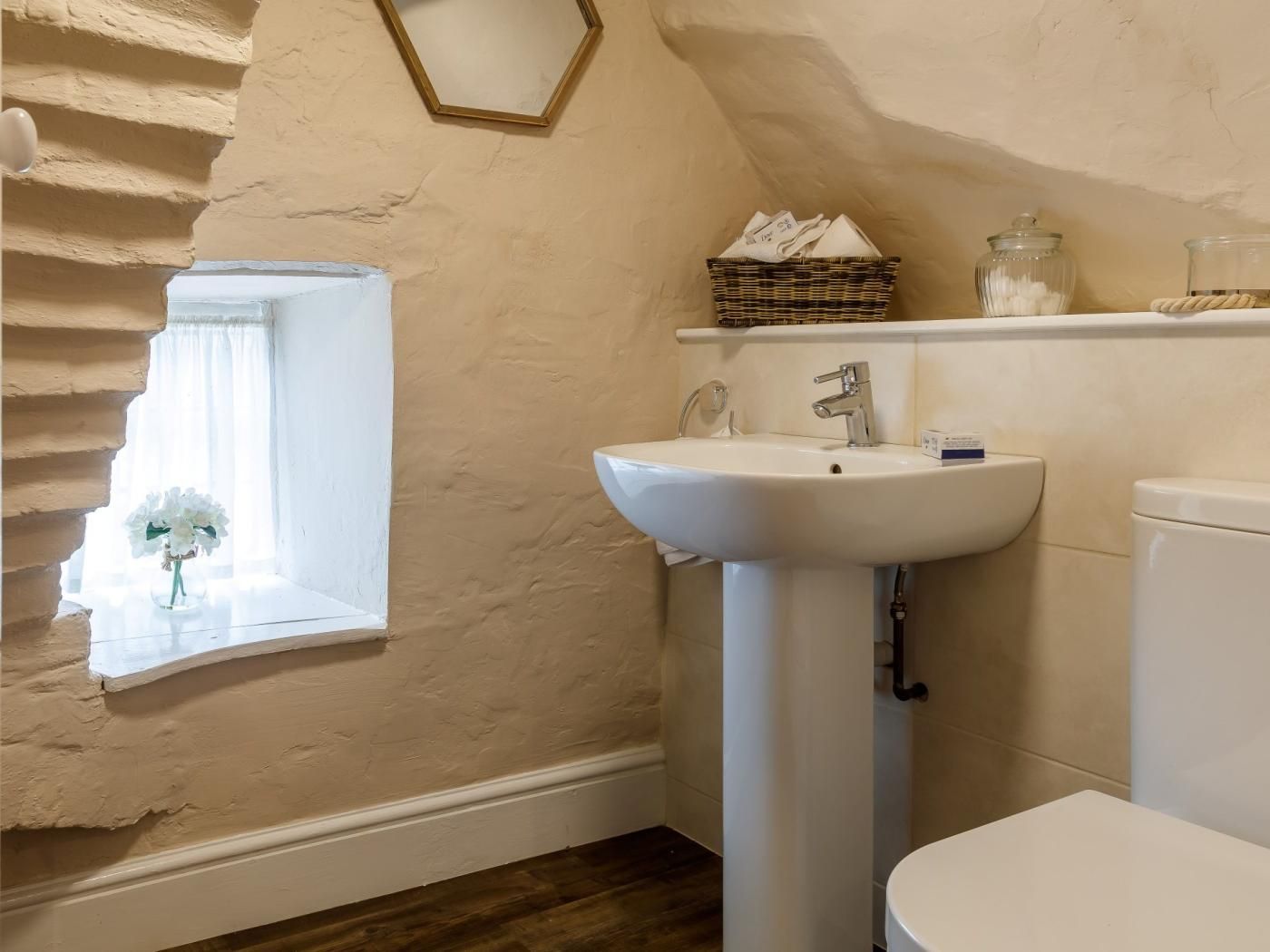 A bathroom with a white sink, a toilet, a window with a white flower arrangement, and a mirror.