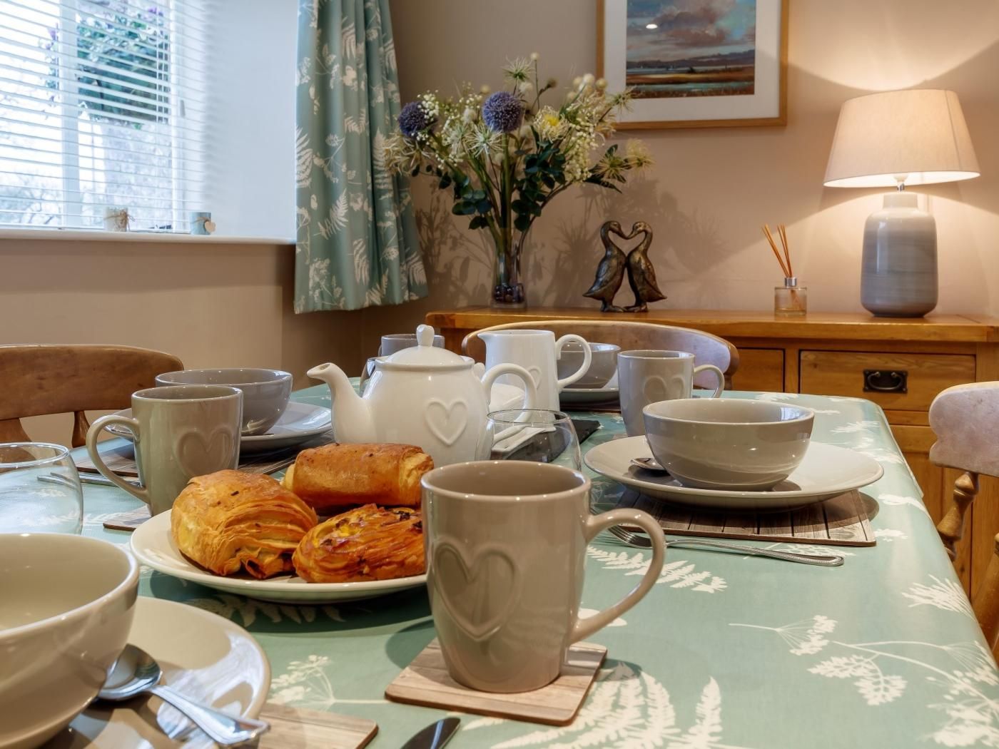 A dining table with a floral tablecloth, heart-shaped mugs, a plate of pastries, and a vase of flowers.
