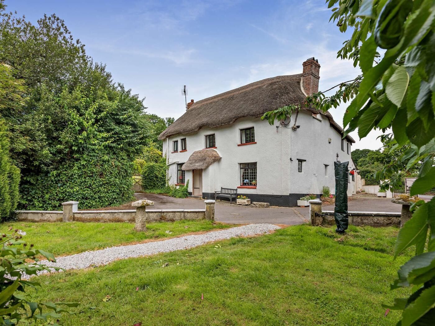 A white building with a thatched roof, surrounded by greenery, and a gravel path leading to the entrance.