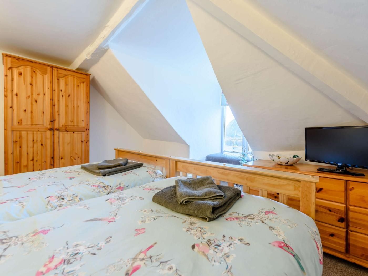 A bedroom with a sloped ceiling, wooden wardrobe, a bed with floral bedding, a bedside table with a TV, and a window.