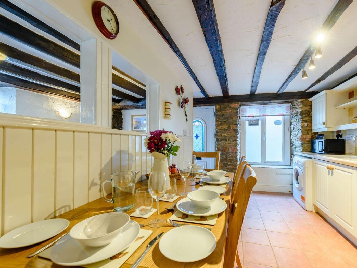A dining area with a wooden table set for four, a vase of flowers, and a clock on the wall.