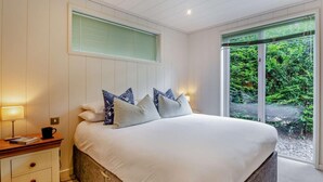 Quarto com cama, mesas de cabeceira, janela com persianas e vista para o verde lá fora.