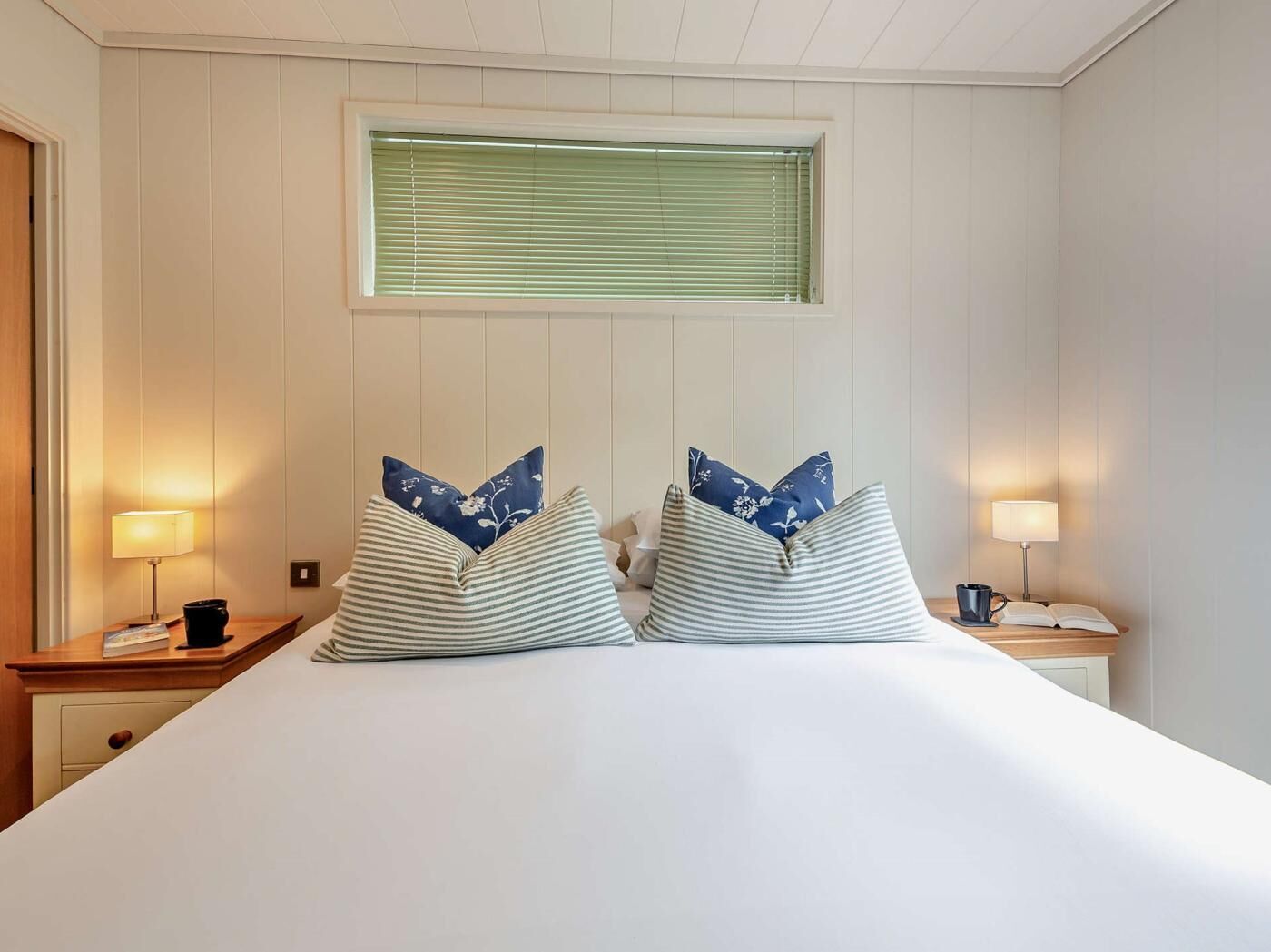 A hotel room with a bed, two bedside lamps, a window with blinds, and a door.