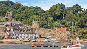 Uma cidade costeira com edifícios de pedra, um porto com barcos e uma colina arborizada ao fundo.