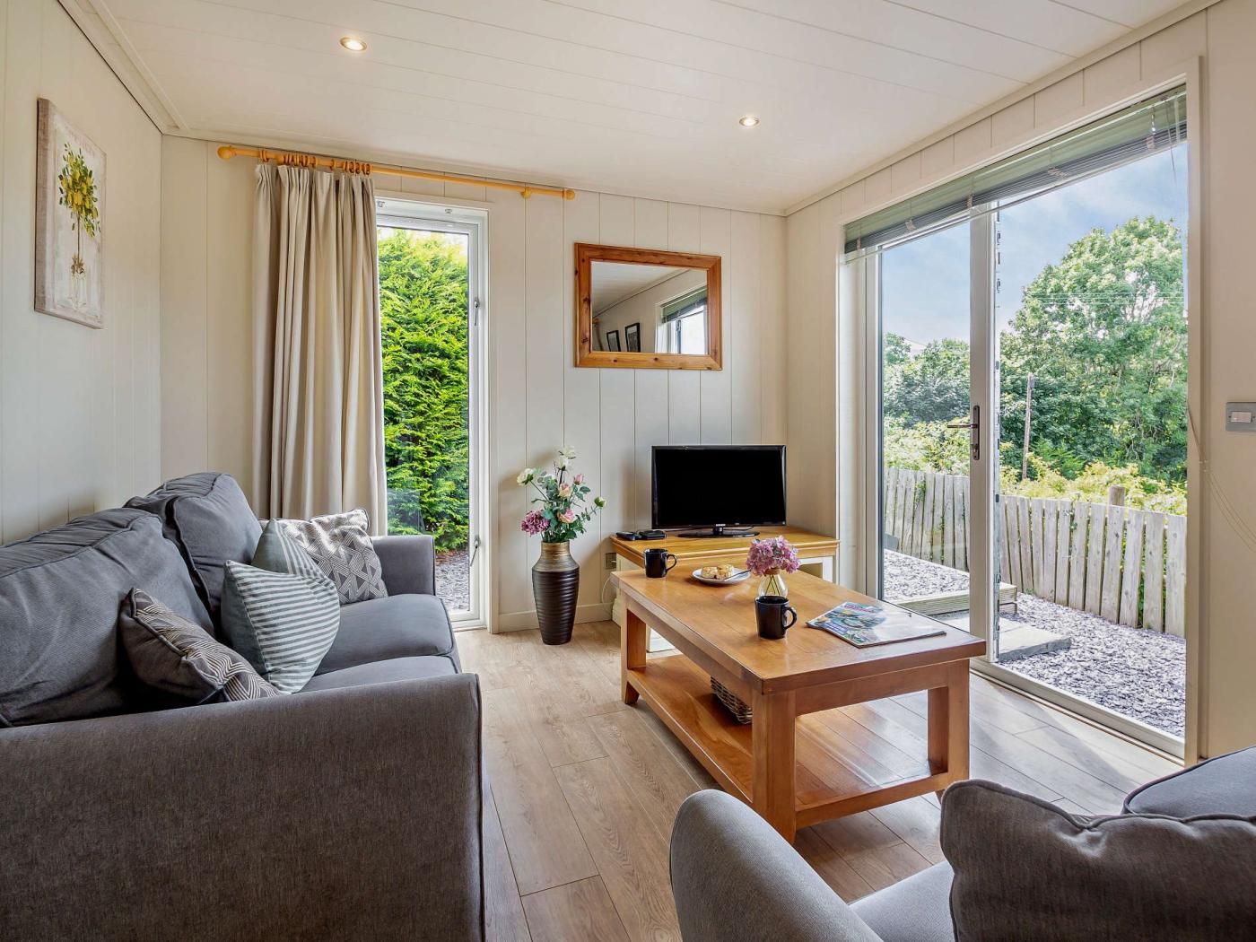 Un salon moderne avec un canapé, une table basse et un téléviseur. Une grande fenêtre offre une vue sur la verdure environnante.