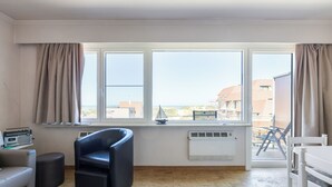 Ein Zimmer mit einem großen Fenster, einem schwarzen Stuhl, einem weißen Stuhl, einem kleinen Tisch und einem Radio.
