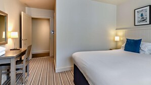 Quarto de hotel com cama, mesas de cabeceira, escrivaninha, cadeira, porta e um quadro na parede.