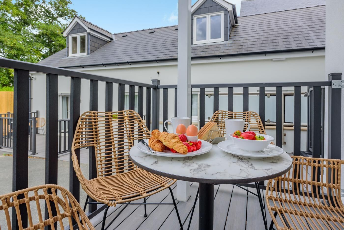 A balcony with wicker chairs, a round table, and breakfast items including croissants, eggs, and strawberries.