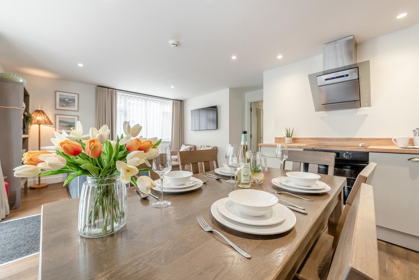 A modern dining area with a wooden table set for a meal, a vase of flowers, and a kitchen in the background.
