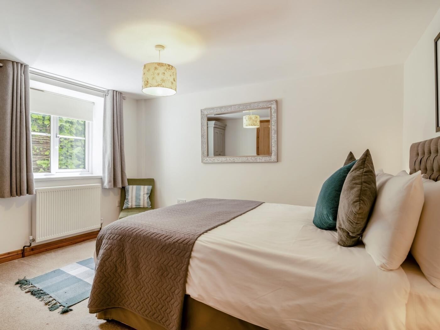 Un dormitorio con cama, ventana con cortinas, radiador, espejo y lámpara colgante.
