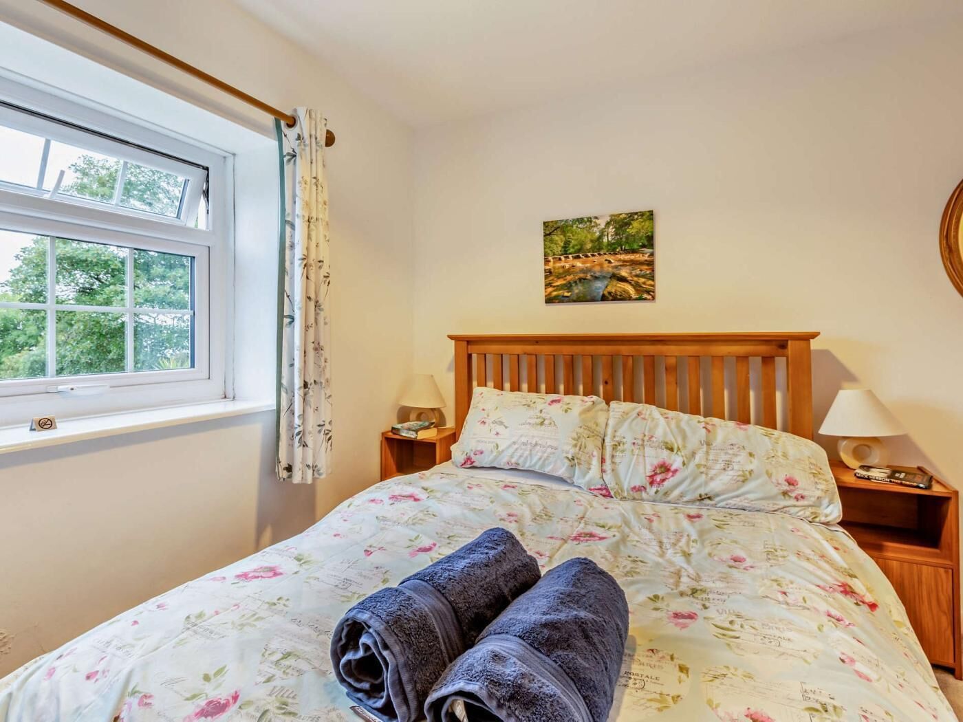 Una cama con ropa de cama floral, una ventana con cortinas, un cuadro en la pared y una lámpara de noche.